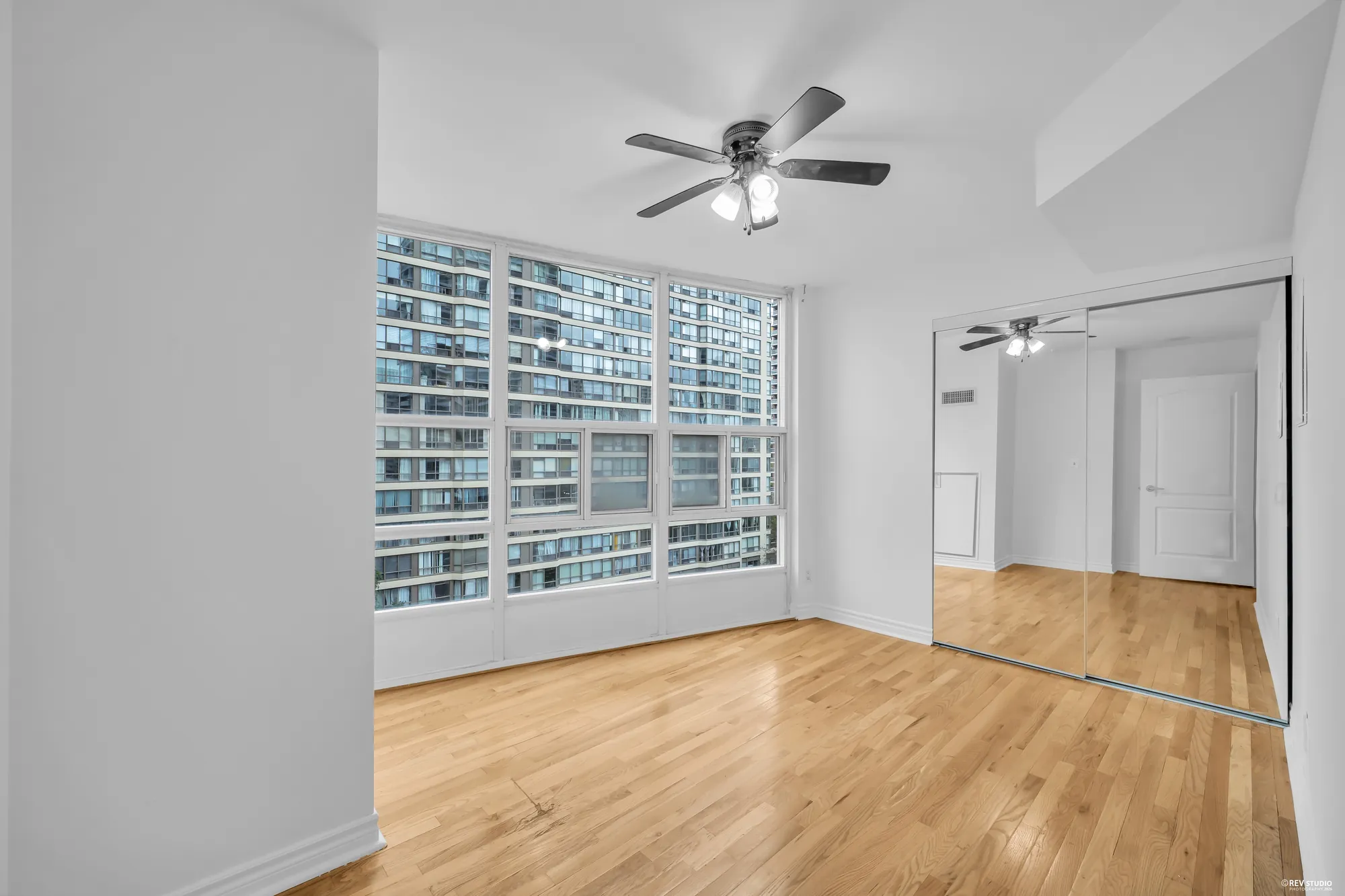 The bedroom, empty: windows on two sides with the towers beyond, a ceiling fan, hardwood, the closet's mirrored doors