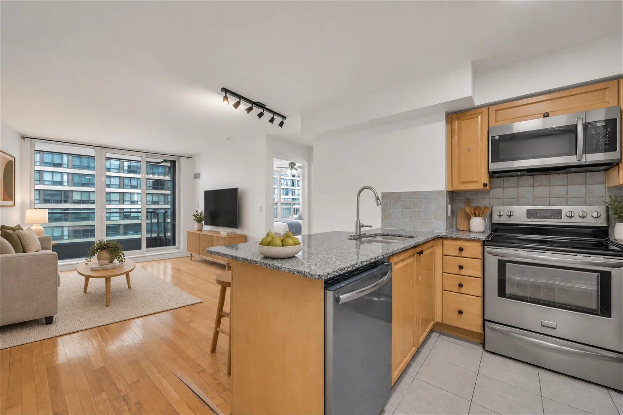 The same kitchen view virtually staged: a fruit bowl and utensils on the counter, stools, and the living room furnished beyond