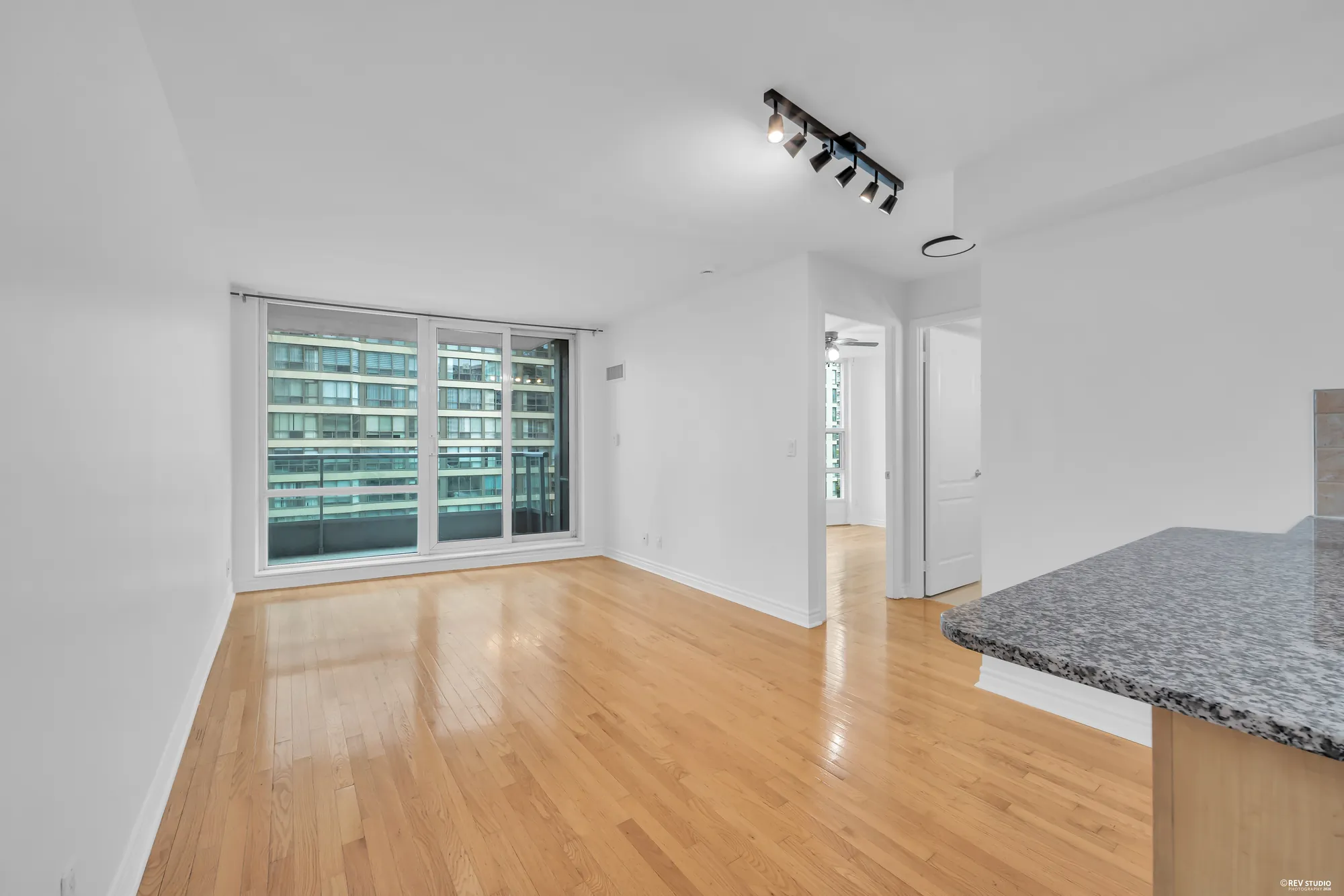 The living and dining room, empty: hardwood floors, white walls, track lighting, a wall of glass to the balcony with the neighbouring tower beyond, the kitchen peninsula's granite counter on the right